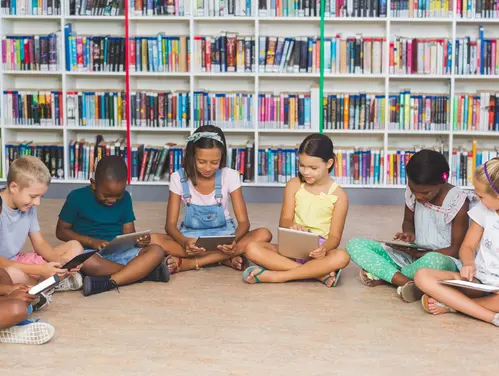 Group of children sitting on the floor of a library and using tablet computers